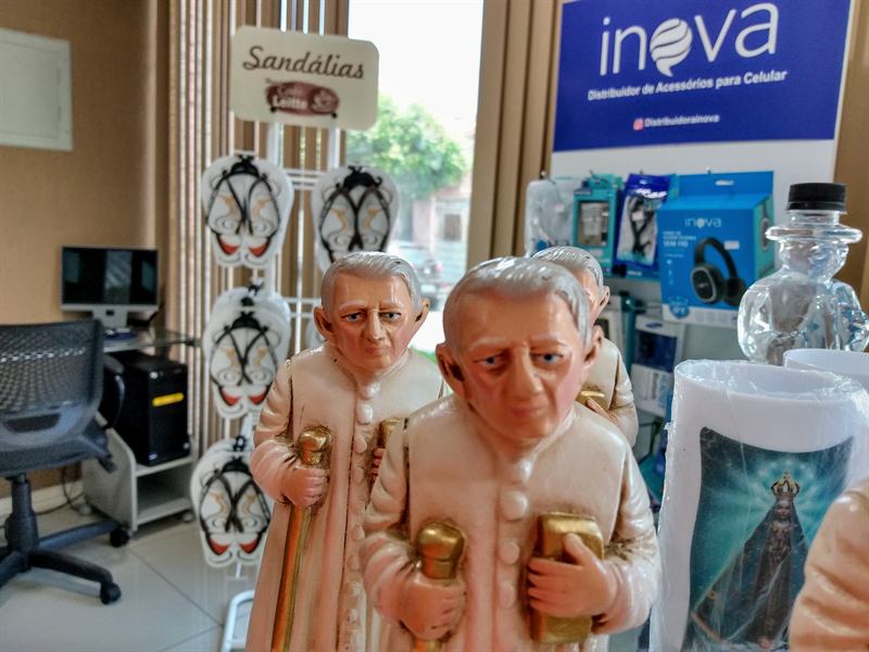 Estatuetas de Padre Cícero em primeiro plano, com o expositor de sandálias e os demais produtos da lojinha ao fundo