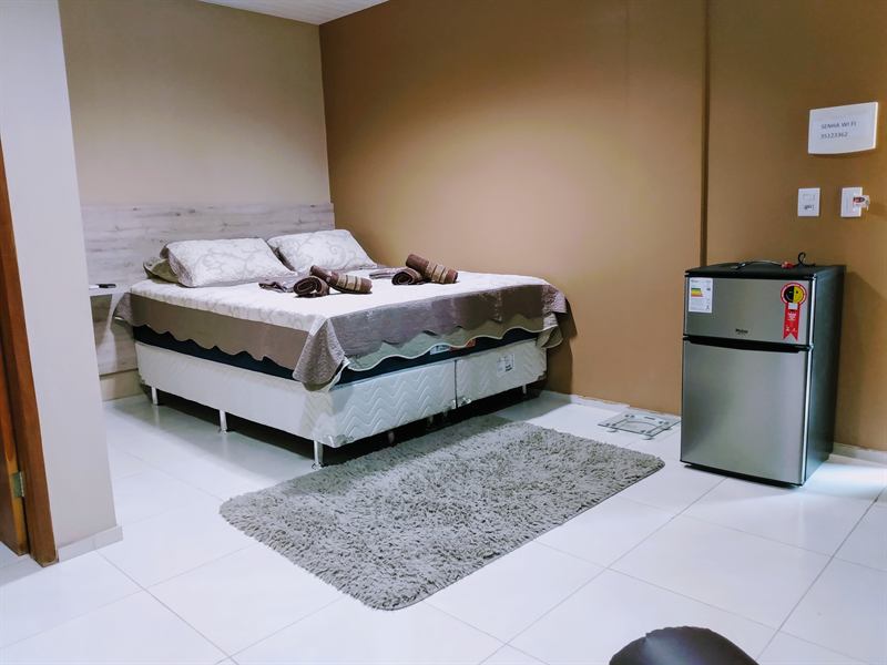 Quarto espaçoso de piso branco com cama de casal arrumada, tapete felpudo e frigobar de aço escovado