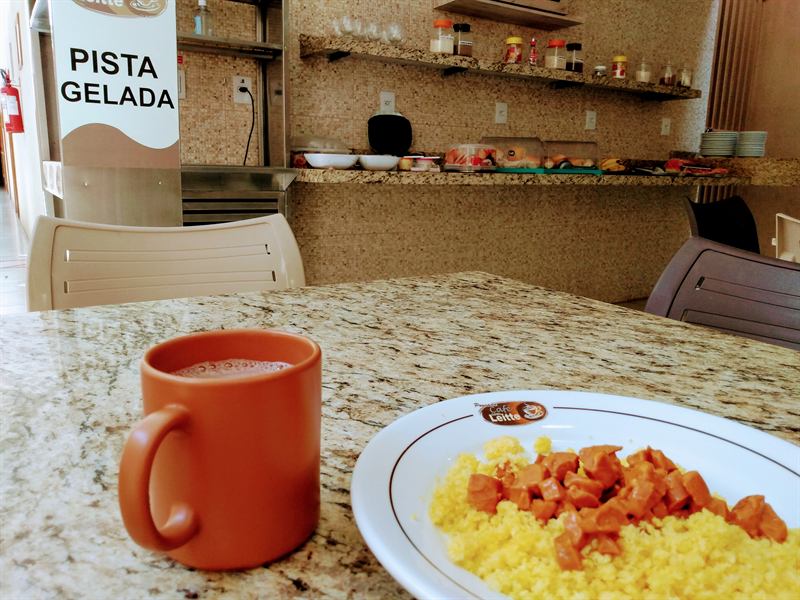 Prato de cuscuz com salsicha ao lado de uma caneca de café sobre a mesa de granito do refeitório