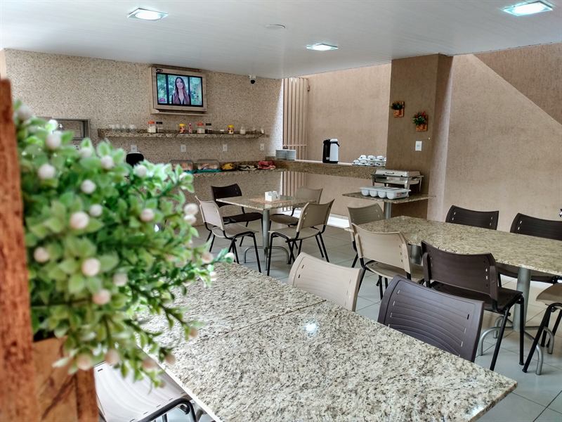 Salão do refeitório com mesas de granito, cadeiras, televisão e a bancada do buffet ao fundo